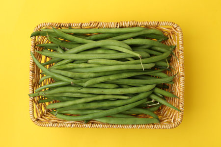 Fresh green bean pods in wicker basket on yellow background, top viewの写真素材