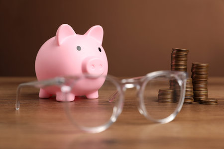 Piggy bank, glasses and coins on wooden table against brown backgroundの写真素材