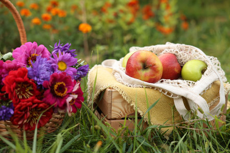 Beautiful bouquet of flowers in wicker basket and apples on green grass outdoors, closeupの写真素材