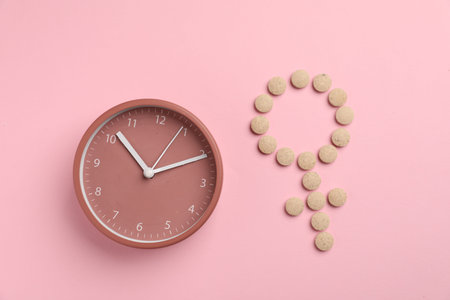 Menopause. Female gender symbol made with pills and alarm clock on pink background, top view