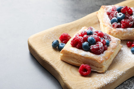 Tasty puff pastries with berries and powdered sugar on gray table, closeup. Space for text