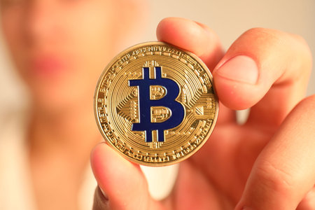 Cryptocurrency. Man holding golden bitcoin against blurred gray background, closeup