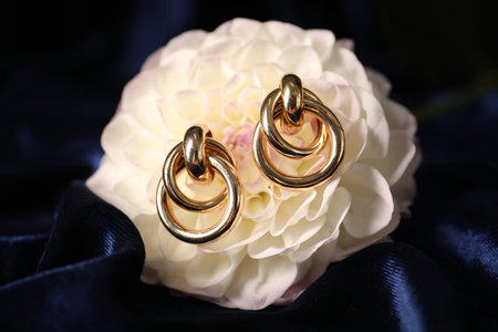 Stylish gold earrings and flower on blue silk cloth, closeup