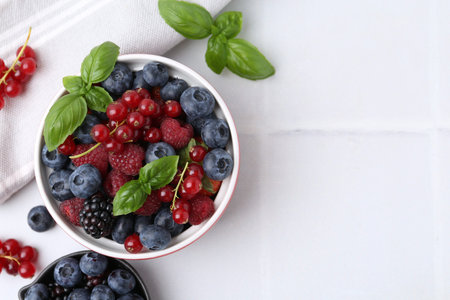 Different ripe berries and basil leaves in bowls on white tiled table, flat lay. Space for text