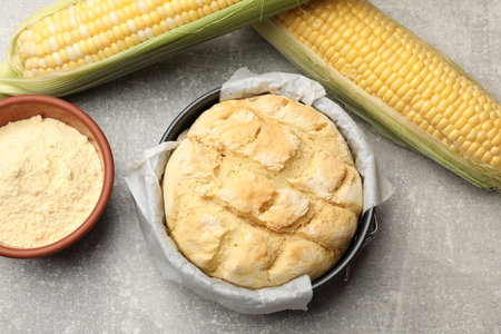 Freshly baked cornbread in cake pan, cobs and flour on gray textured table, flat lay