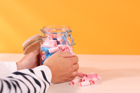 Woman with glass jar of color paper pieces at white wooden table, closeup. Space for textの写真素材