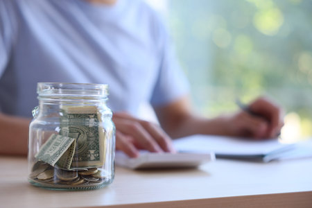 Glass jar with money and man budgeting at wooden desk, selective focus