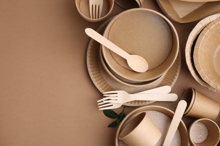 Set of disposable tableware and green leaves on brown background, flat lay. Space for text