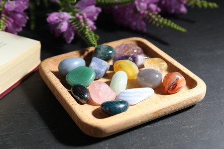Different beautiful gemstones, book and flowers on black table, closeup