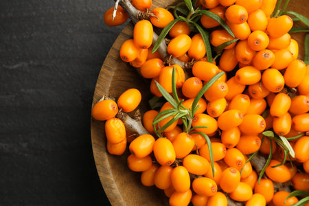 Fresh sea buckthorn berries with leaves in bowl on black table, top view