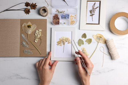 Woman putting dry flowers into picture frame at white marble table, top view