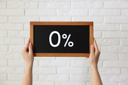 Woman holding chalkboard with 0% against white brick wall, closeup
