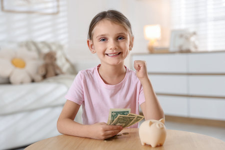 Little girl with pocket money and piggy bank at wooden table indoorsの写真素材