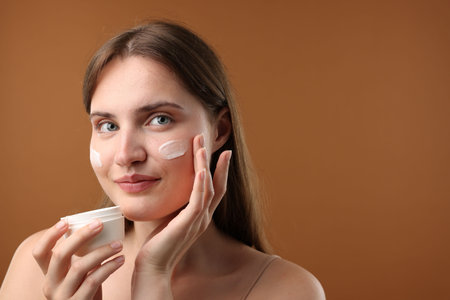 Beautiful woman applying cream onto her face on brown background. Space for textの写真素材
