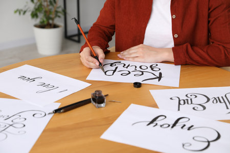 Calligraphy. Woman with brush writing phrase on sheet of paper at wooden table indoors, closeupの写真素材