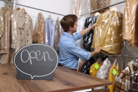 Dry-cleaning service. Worker choosing clothes from rack, focus on Open signの写真素材
