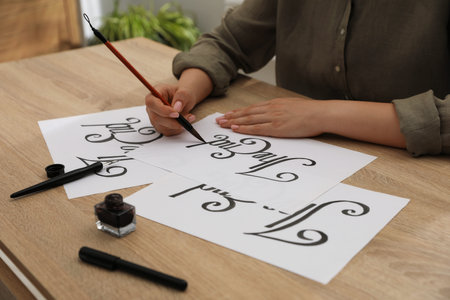 Calligraphy. Woman with brush writing words The End on sheet of paper at wooden table, closeup