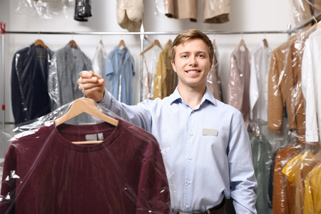 Dry-cleaning service. Happy worker holding jacket in plastic bag indoorsの写真素材