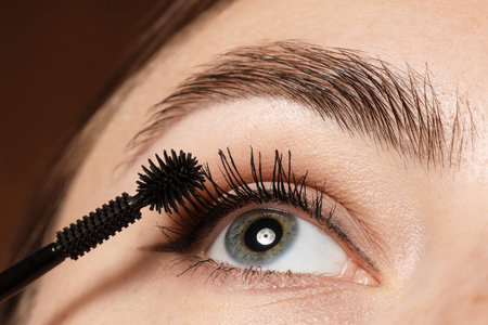 Woman applying mascara onto her eyelashes on brown background, closeup