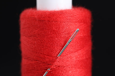 Spool of red thread with sewing needle on black background, macro view