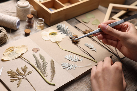 Woman putting dry flowers into notebook at wooden table, closeup