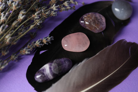 Different beautiful gemstones in bowl, feathers and lavender flowers on purple background, closeup