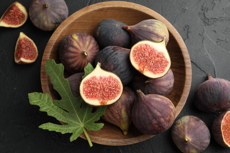 Fresh ripe figs in bowl and green leaf on black textured table, flat lay