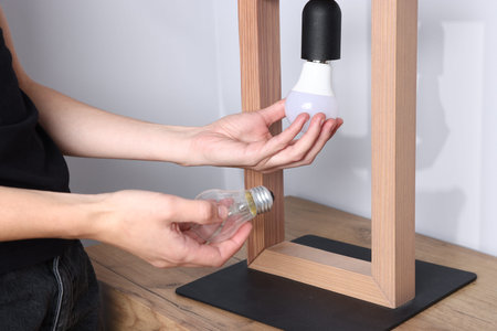 Woman changing light bulb in lamp near gray wall at wooden chest of drawers indoors, closeup