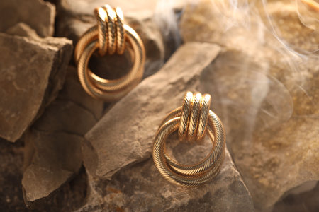 Stylish gold earrings in smoke on rocks, closeup