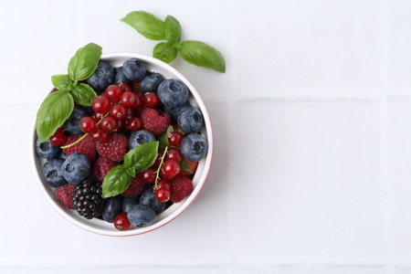 Different ripe berries and basil leaves in bowl on white tiled table, flat lay. Space for text