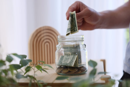Man putting money into jar at wooden table indoors, closeup