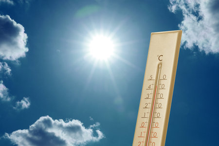Thermometer against blue sky with clouds and bright sun. Extremely hot weather