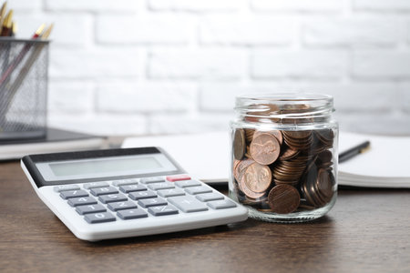 Glass jar with coins and calculator on wooden table, closeup