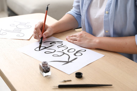 Calligraphy. Woman with brush writing phrase on sheet of paper at wooden table indoors, closeupの写真素材