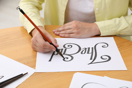 Calligraphy. Woman with brush writing words on sheet of paper at wooden table indoors, closeup