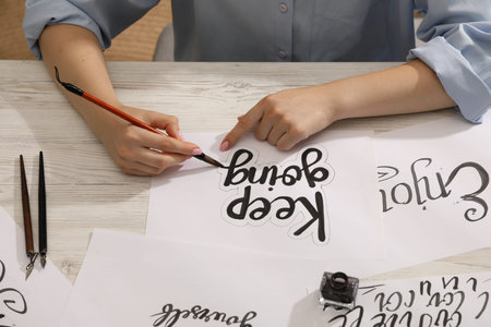 Calligraphy. Woman with brush writing phrase Keep Going on sheet of paper at wooden table, closeup