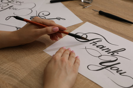 Calligraphy. Woman with brush writing words Thank You on sheet of paper at wooden table, closeupの写真素材