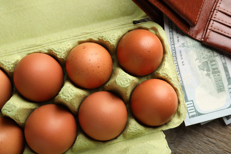 Raw chicken eggs in egg carton and dollar banknotes on wooden table, above view