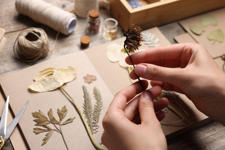 Woman putting dry flowers into notebook at wooden table, closeup