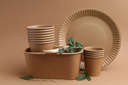 Set of disposable tableware and green leaves on brown background