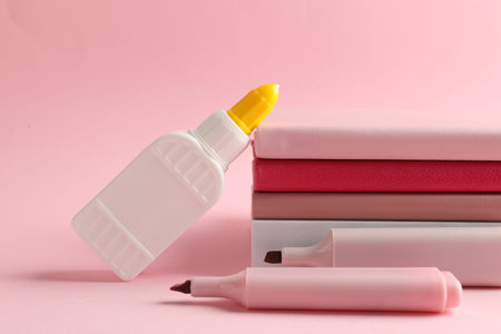 Glue in bottle, felt pens and notebooks on pink background, closeup