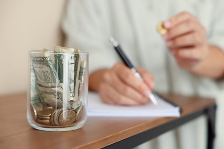 Woman putting money into jar and writing in notebook at wooden table indoors, selective focus