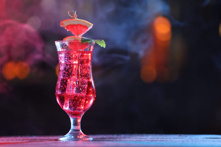 Glass of alcoholic cocktail with garnish on table against blurred background with smoke, space for...