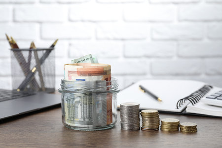 Glass jar with banknotes, coins, notebook and laptop on wooden table, closeup