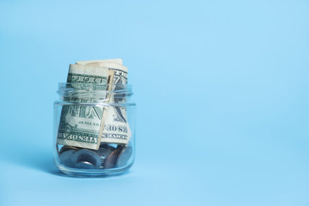 Glass jar with coins and dollars on light blue background, closeup. Space for text