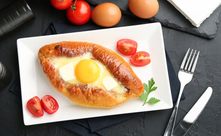Tasty khachapuri with cheese and egg served on black textured table, flat lay