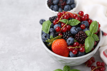 Different ripe berries and basil leaves in bowls on light gray textured table, closeup. Space for textの写真素材