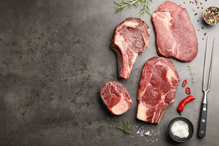 Pieces of raw beef meat, spices and carving fork on gray table, flat lay. Space for text