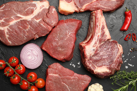 Pieces of raw beef meat, spices and tomatoes on black table, flat lay