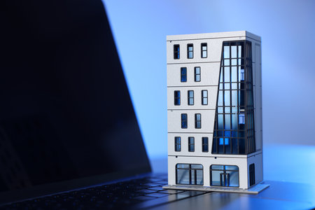 Real estate. Apartment building model and laptop on table in color light, closeup